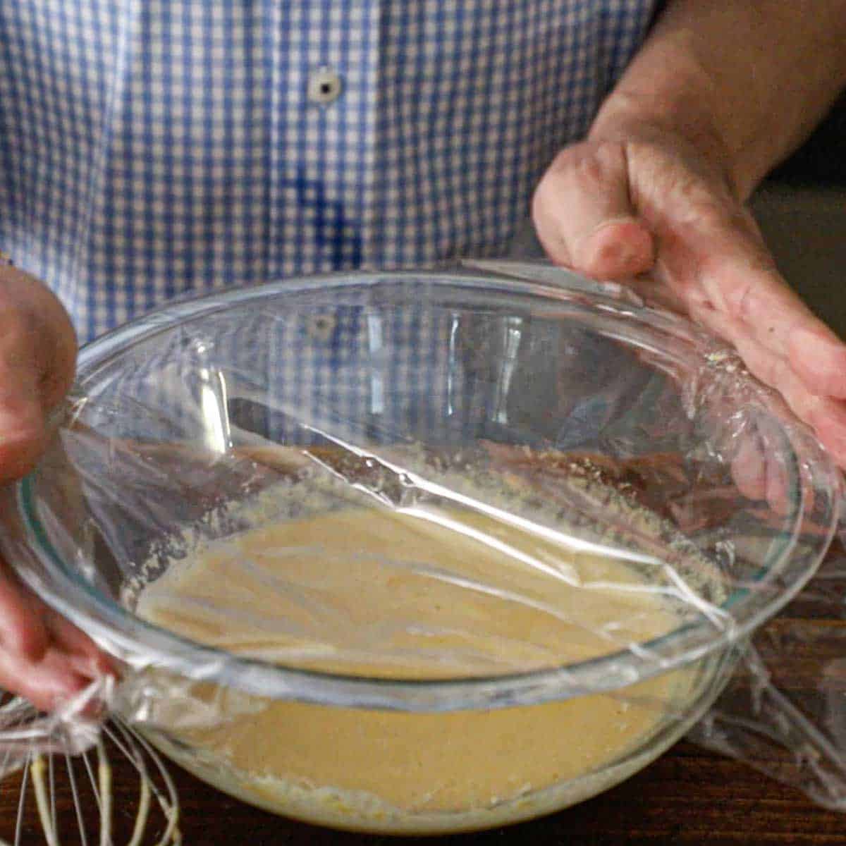 A person placing plastic wrap over a glass bowl that contains batter for easy homemade socca on a wooden cutting board.