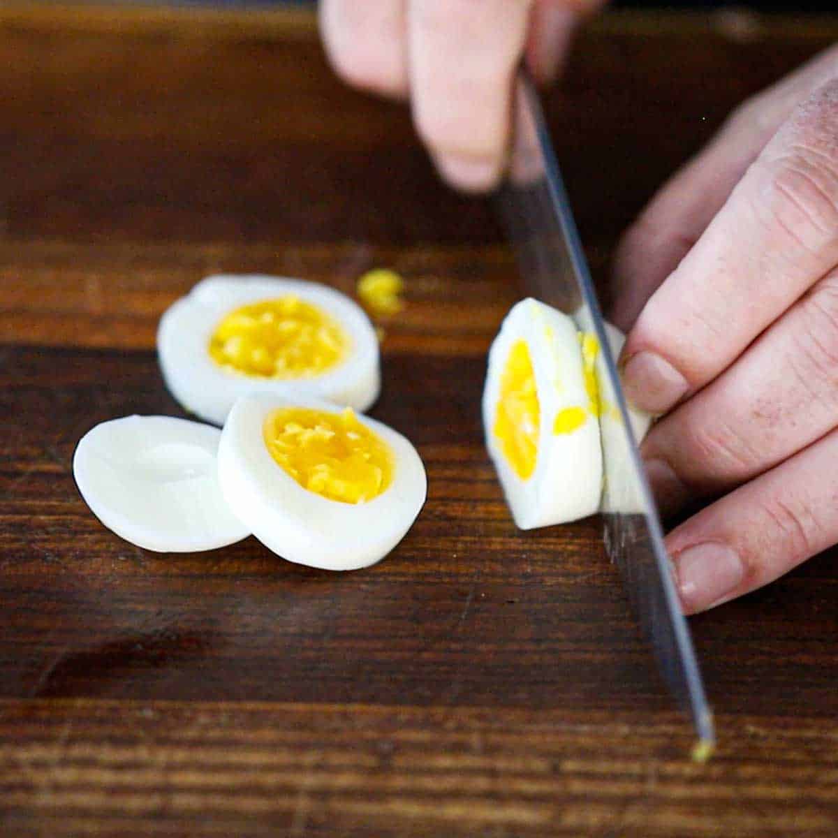 A person using a chef's knife to slice a soft-boiled egg into slices on a wooden cutting board.