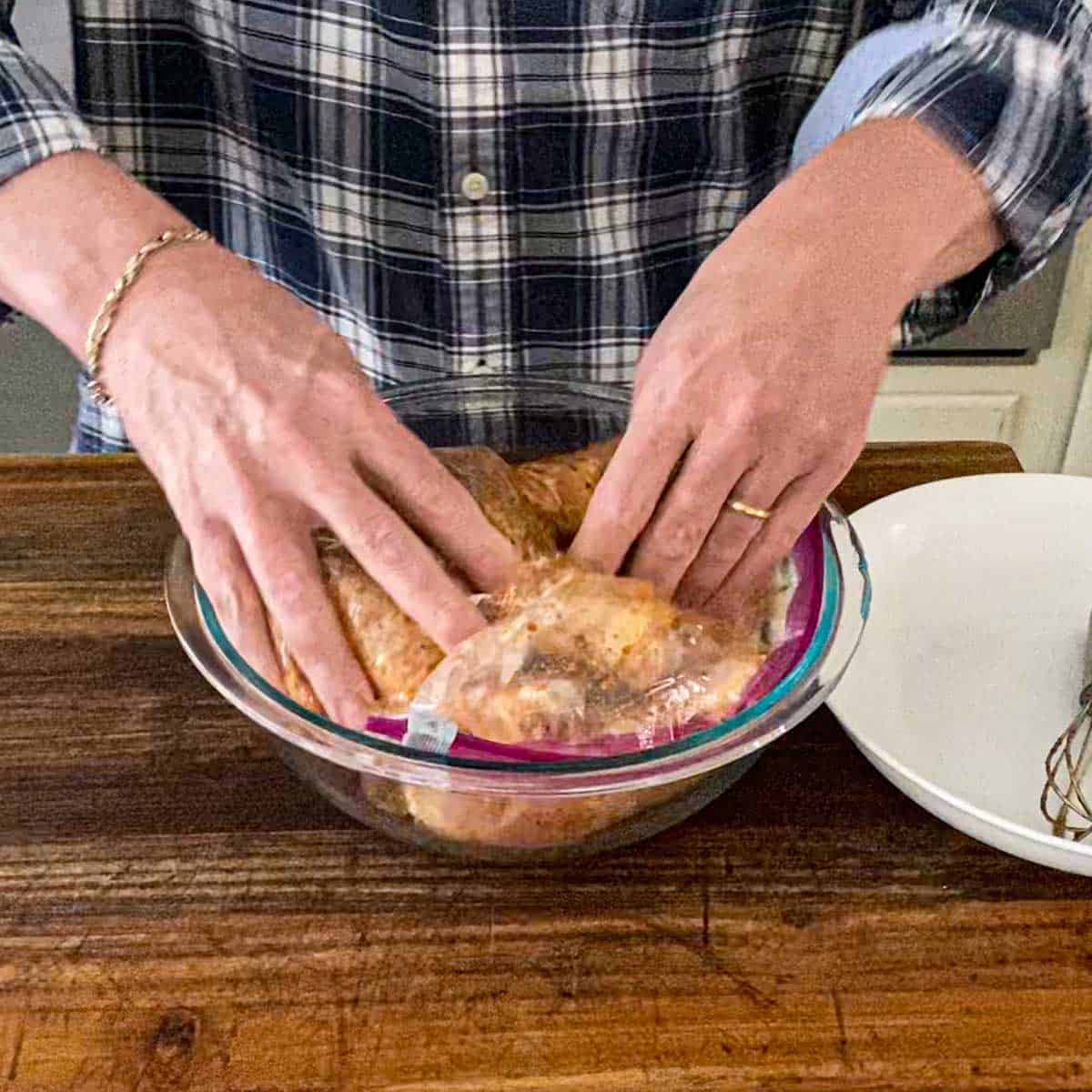 A person using his hands to massage a Peruvian marinade into chicken pieces that are inside a large freezer baggie resting in a large glass bowl.