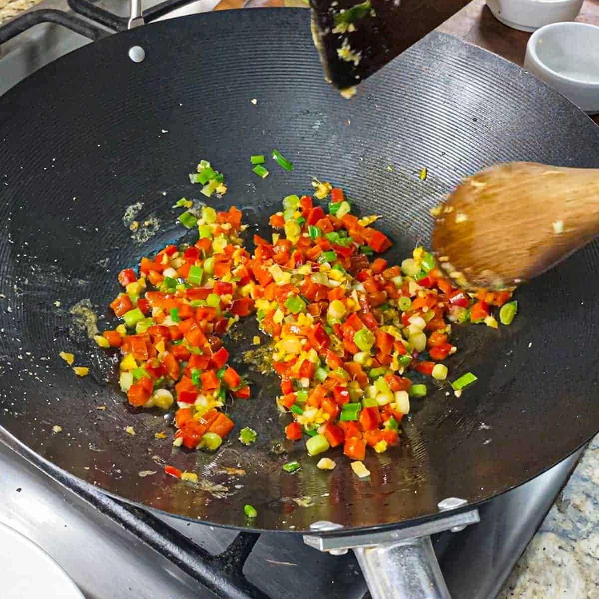A person using two wooden spoons to stir-fry diced red bell pepper, chopped scallions, and minced ginger in oil in a wok over a gas stove.