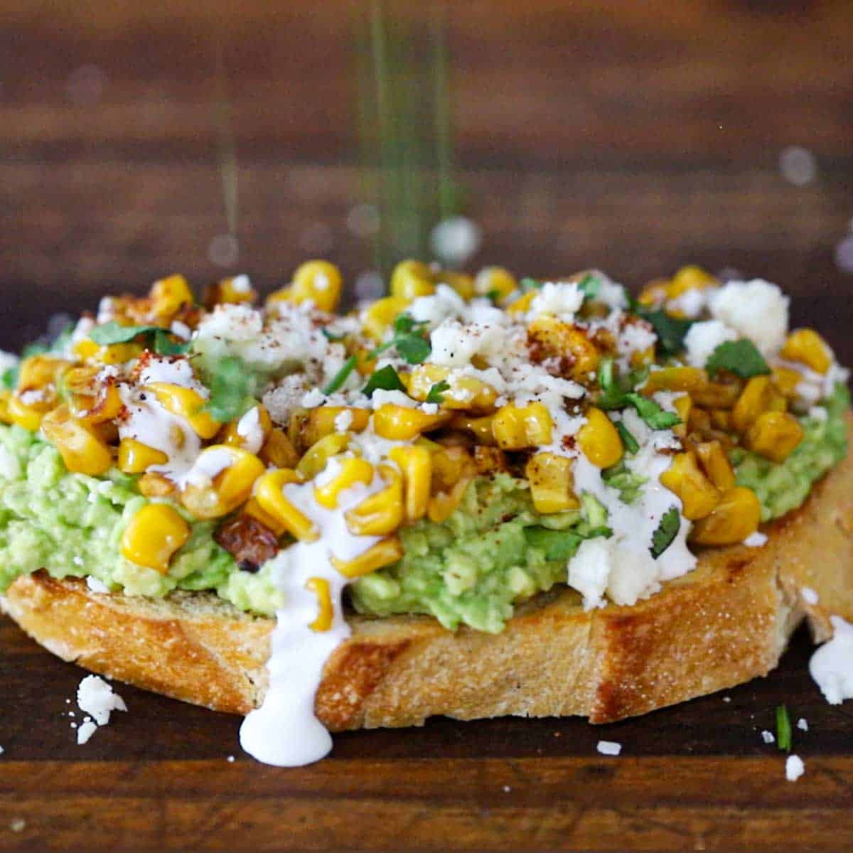 A close-up view of an avocado toast that is topped with roasted corn, Mexican crema, crumbled Cotija cheese, chopped cilantro, and is being dusted with chili powder.