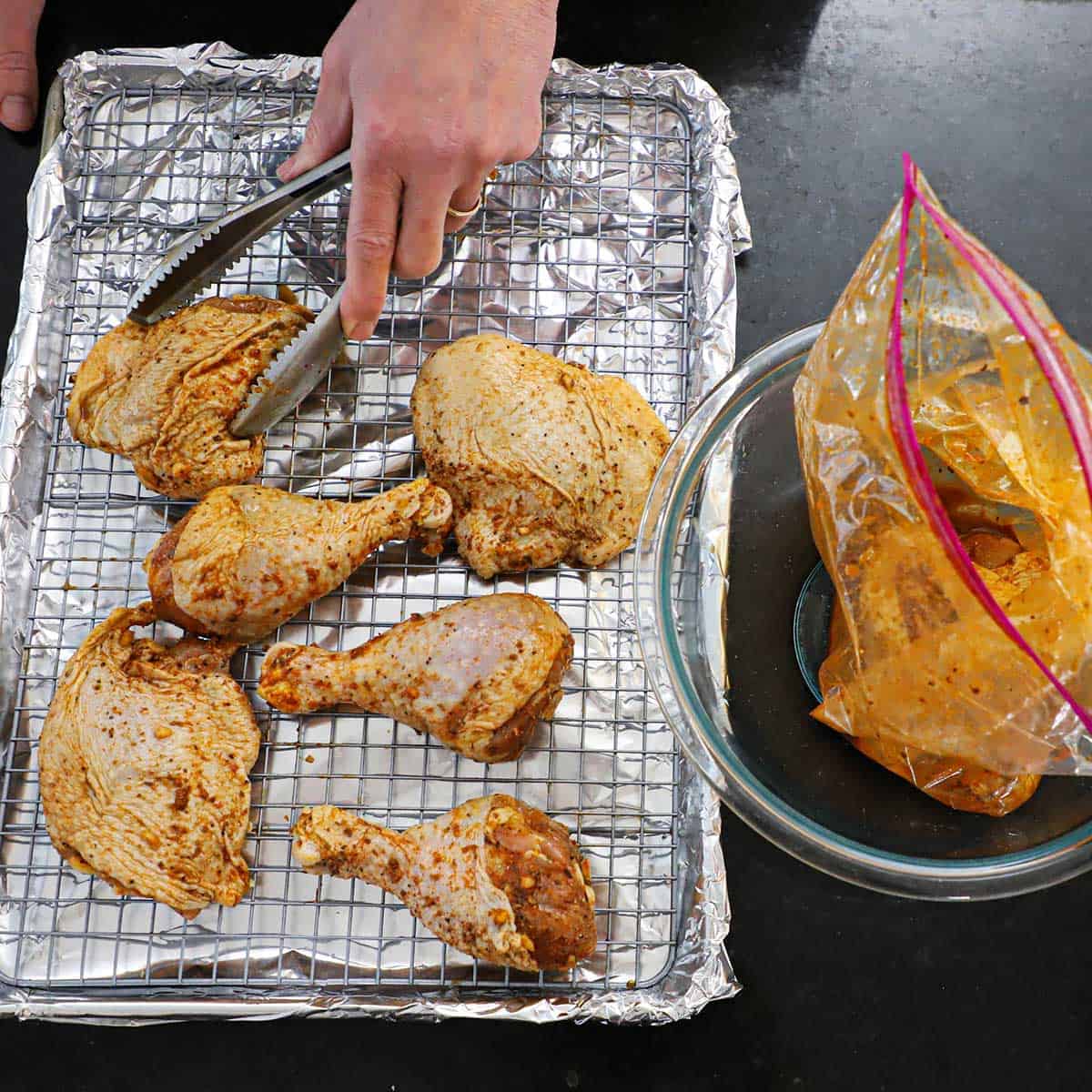 A person using a pair of tongs to place marinated chicken legs and thighs onto a baking rack that is resting inside a foil-lined baking sheet pan.