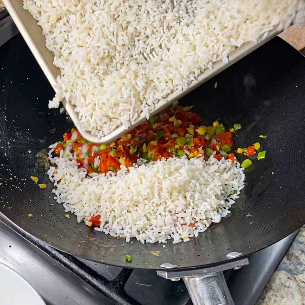 A person transferring day-old cooked white rice from a baking pan into a wok filled with simmering chopped vegetables in vegetable oil.