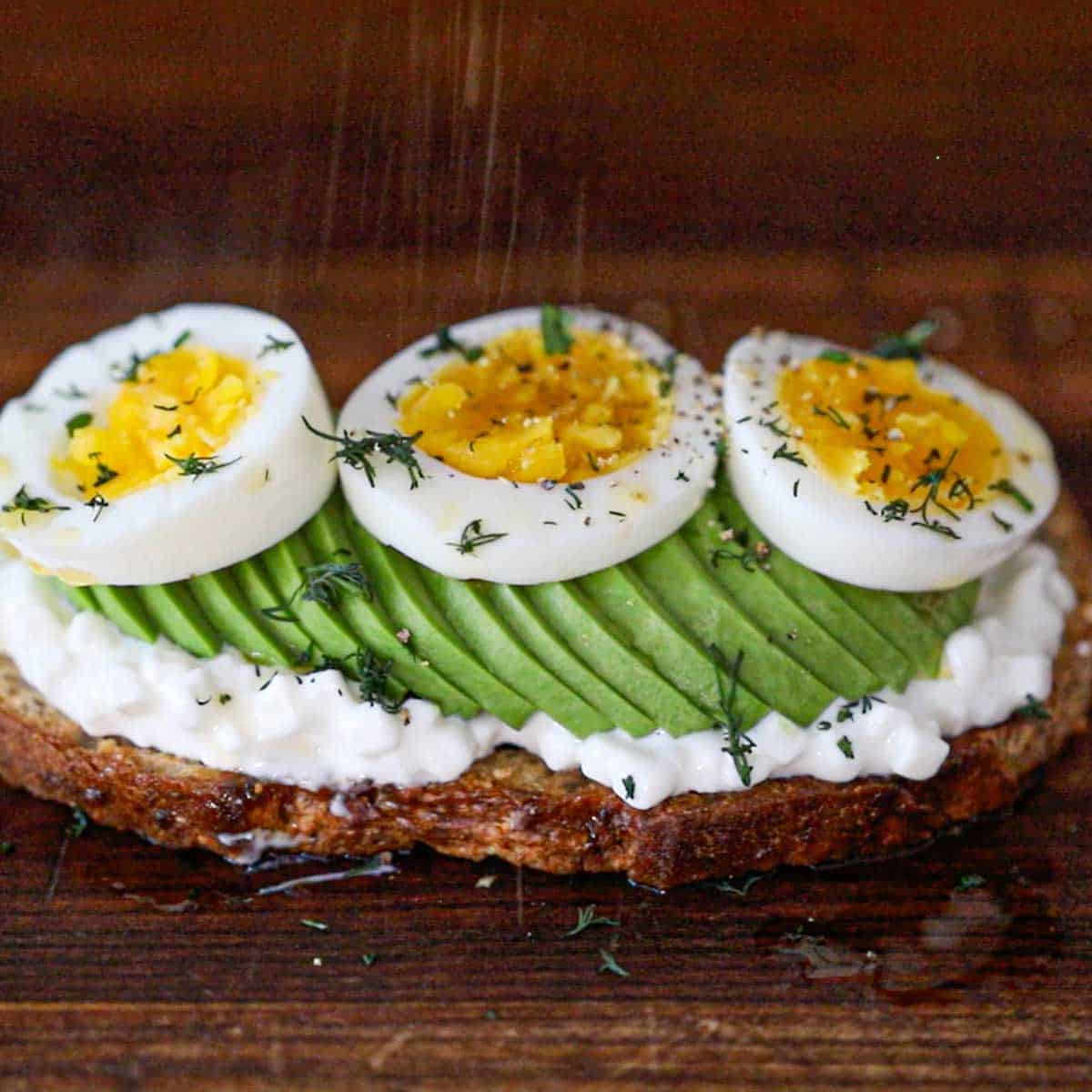 A close-up view of an egg and cottage cheese avocado toast that is topped with chopped dill and is being sprinkled with salt and pepper.