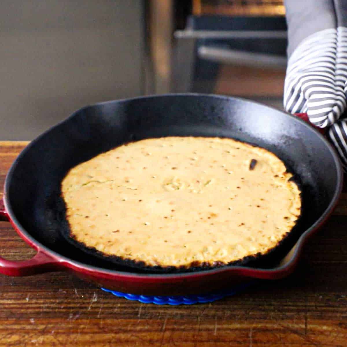 A person wearing oven mitts placing an extremely hot skillet with freshly baked easy homemade socca in down onto a hot pad on a wooden cutting board.