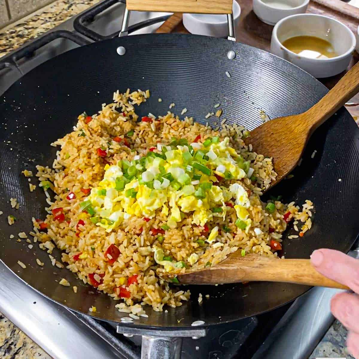 A person using two large wooden spoons to toss cooked, chopped scrambled eggs and chopped scallions into Peruvian fried rice that is being cooked in a large wok on a gas stove.
