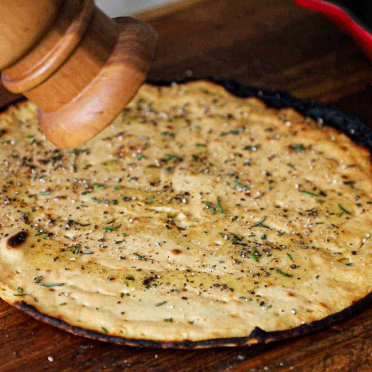 A person adding ground black pepper from a pepper grinder onto a freshly baked easy homemade socca on a wooden cutting board.