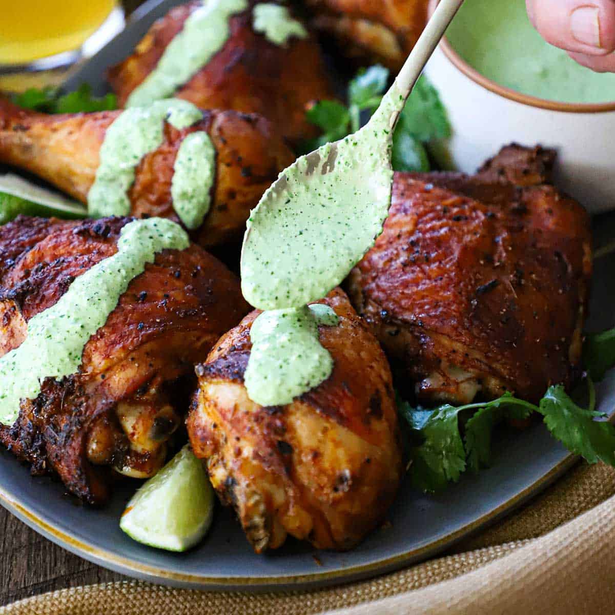 A person using a spoon to drizzle a vibrant green sauce of the top roasted Peruvian chicken pieces on a grey platter.