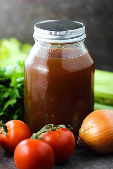 A large glass jar with a metal lid holding homemade beef stock with several tomatoes, an onion, herbs, and celery arranged around it.