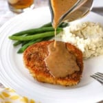 A person pouring brown gravy from a glass gravy boat over the top of a crispy pan-fried pork chop on a white dinner plate also containing a pile of mashed potatoes and steamed green beans.