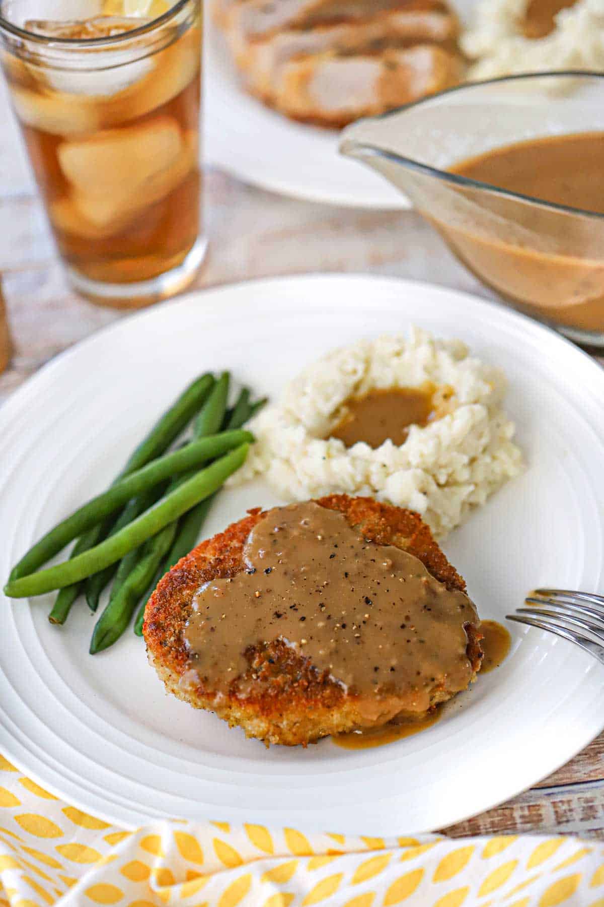 A white dinner plate filled with a serving of Crispy Pan-Fried Pork Chops and Gravy along with mashed potatoes with gravy, and a small serving of steam green beans.