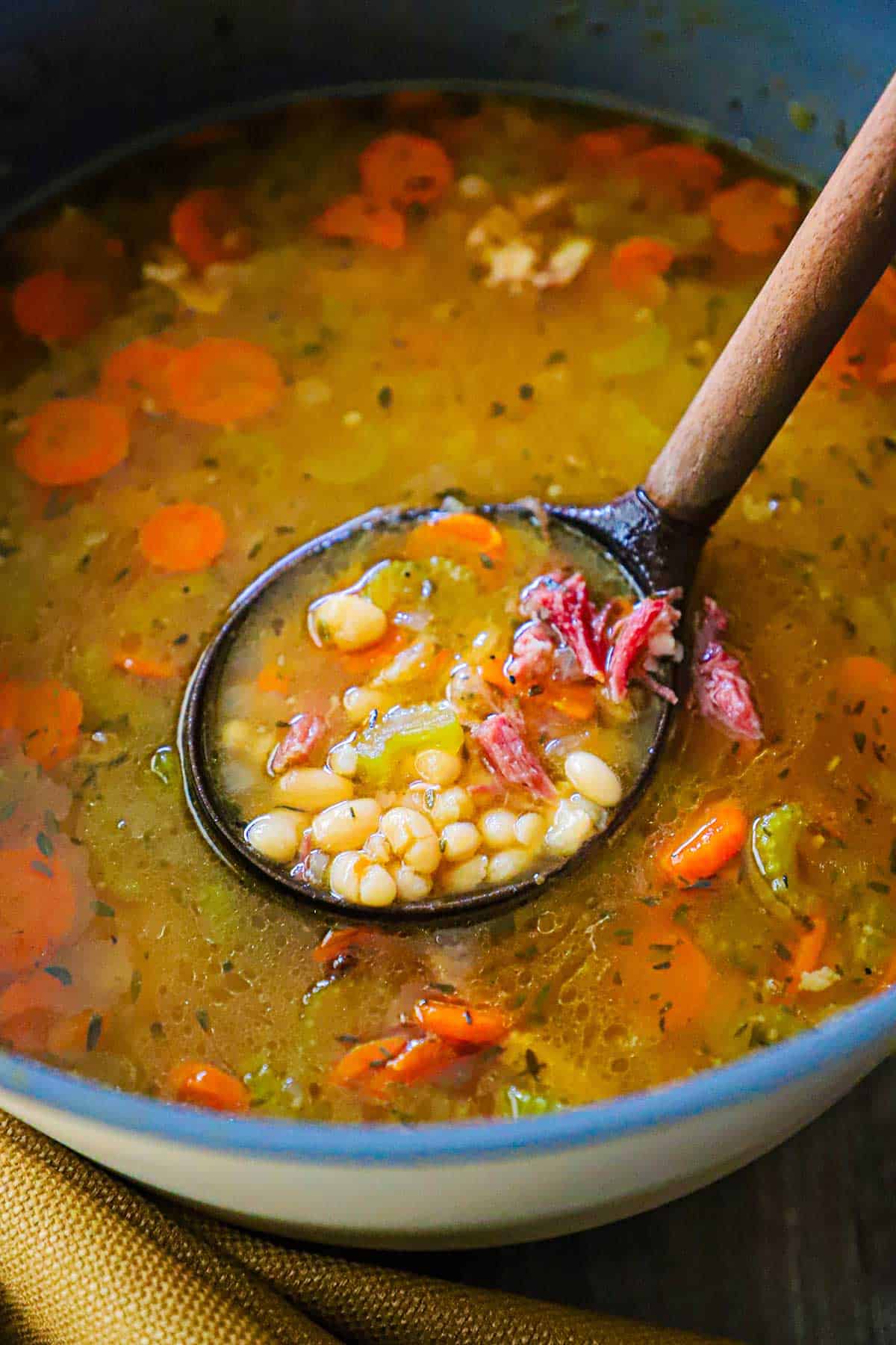 An overhead view of a large stock pot filled with Classic Ham and Navy Bean Soup with a wooden ladle inserted in the middle of it lifting a serving of it.