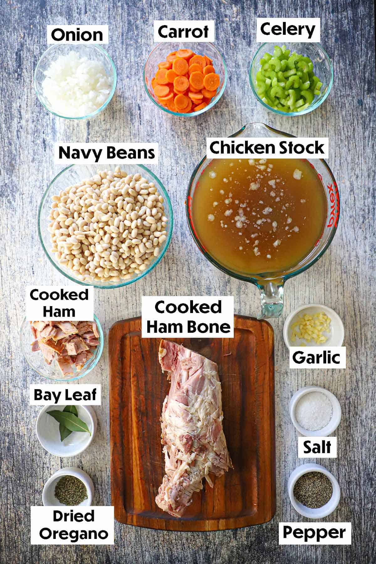 An arrangement of ingredients for Classic Ham and Bean Soup on a grey wooden background including a cooked ham bone, navy beans, onion, carrots, celery, chicken stock, ham, garlic, and seasonings.