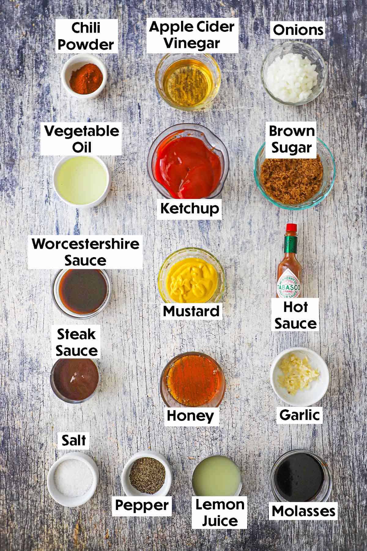 An arrangement of ingredients for homemade easy BBQ sauce on a wooden grey background including ketchup, mustard, vinegar, onions, brown sugar, Worcestershire sauce, garlic, steak sauce, honey, lemon juice, and seasonings.