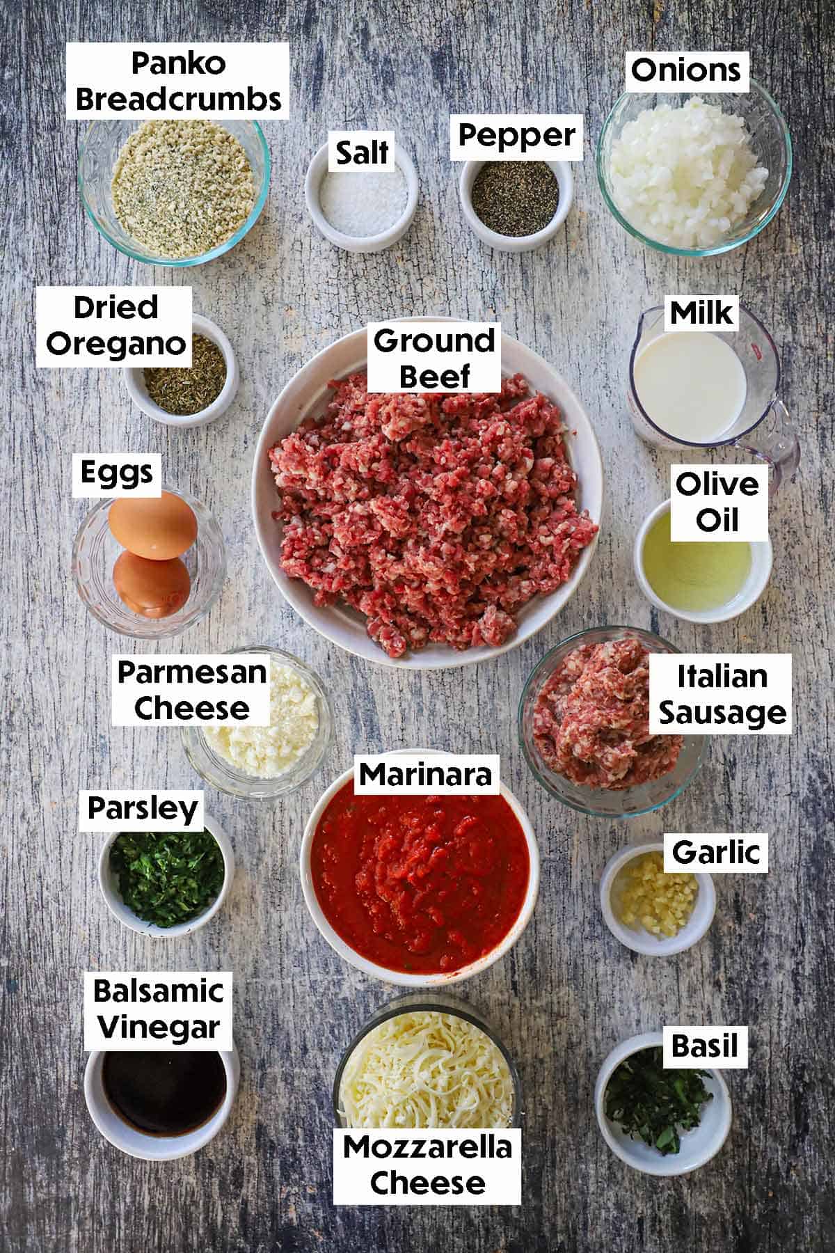 An arrangement of ingredients for Italian-style meatloaf on a grey wooden background including ground beef, Italian sausage, breadcrumbs, milk, onion, garlic, eggs, marinara sauce, balsamic vinegar, herbs, and spices.