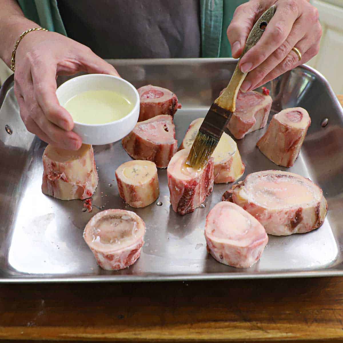 A person using a brush to apply vegetable oil to cut beef marrow bones on a large steel roasting pan.