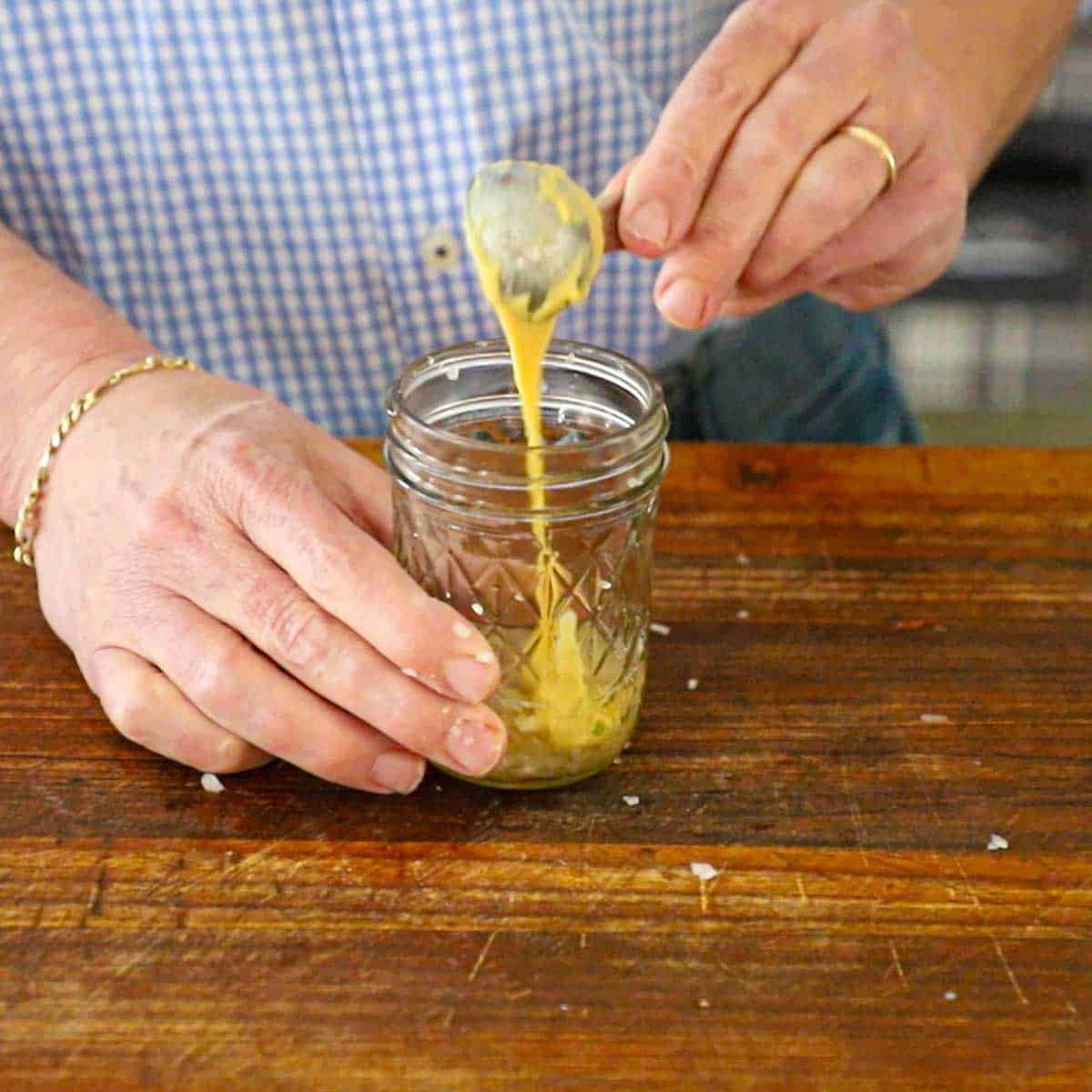 A person transferring a tablespoon of Dijon mustard into a small jar filled with finely chopped shallots and white wine vinegar.