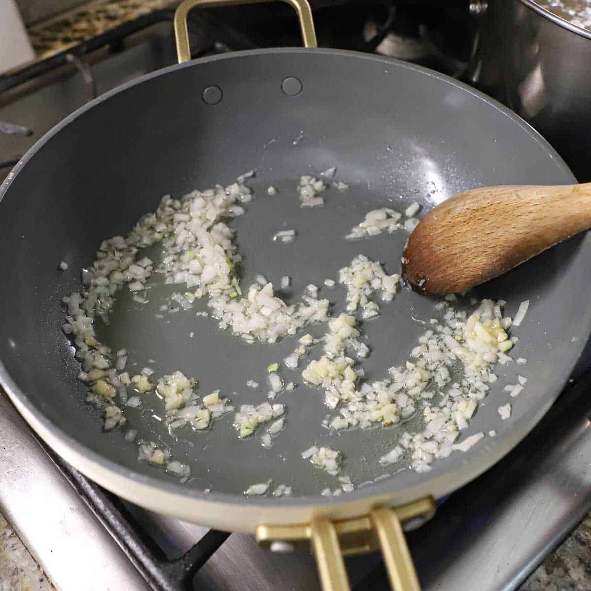A person using a wooden spoon to stir and sauté a chopped shallot in olive oil in a non-stick skillet on a gas stove.