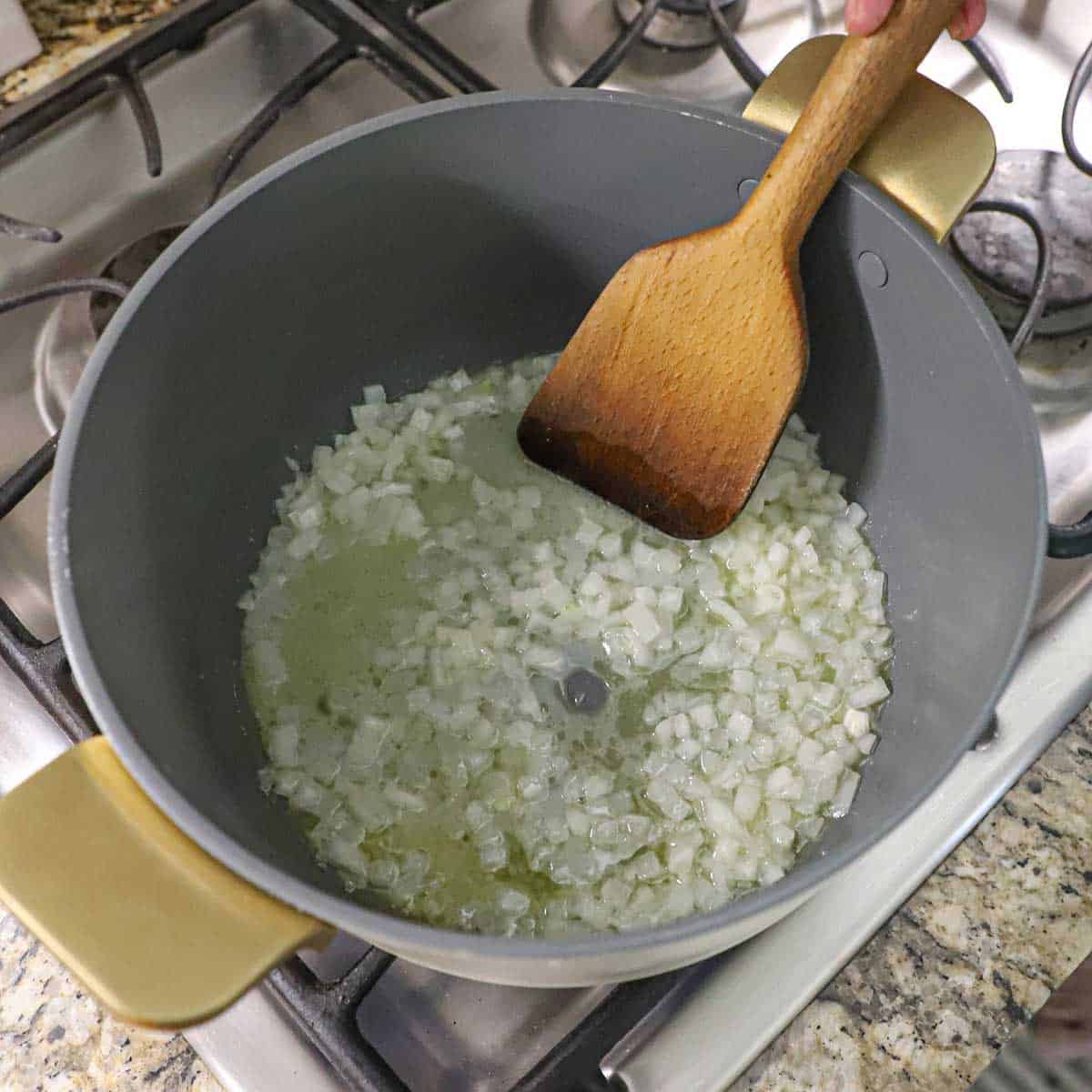A person using a wooden spatula to stir and sauté chopped onions in a large pot on a gas stove.