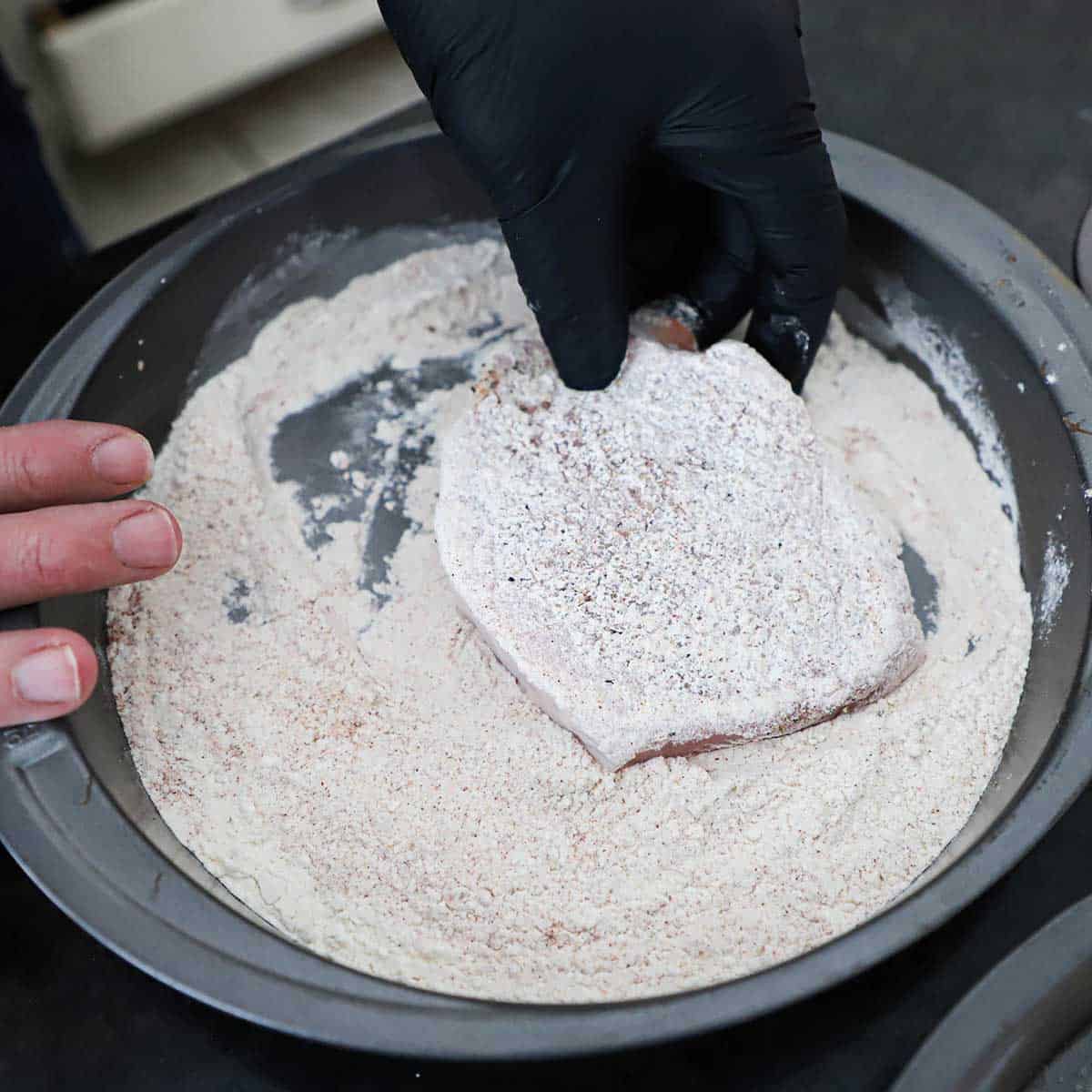 A person wearing a black latex glove dredging a boneless pork chop through seasoned flour that is in a round baking pan.