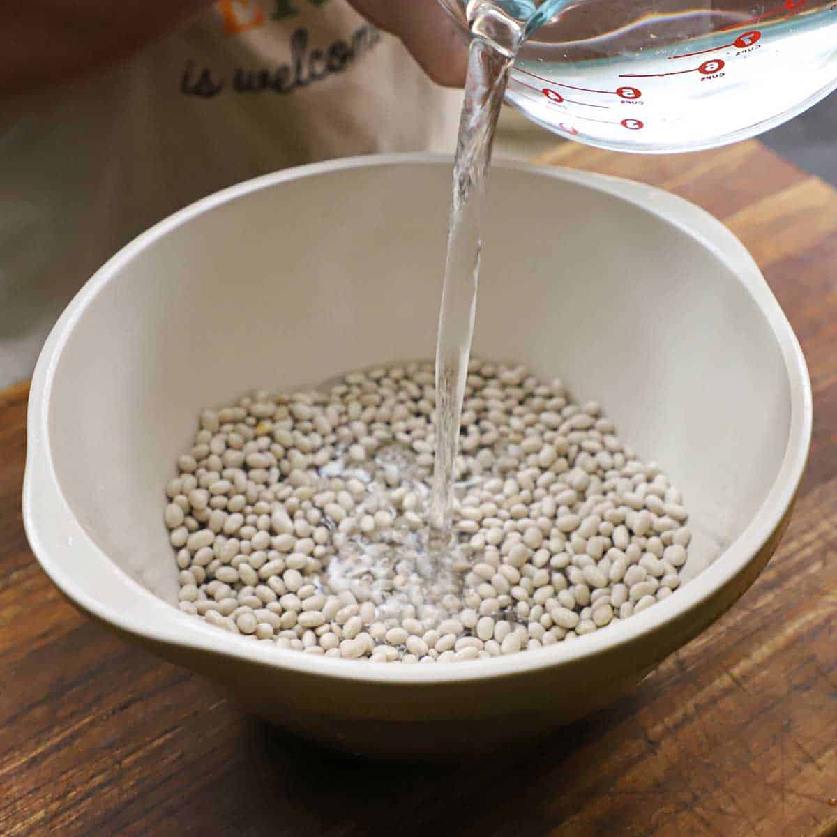 A person pouring water from a large glass measuring cup into a white ceramic bowl filled with dried navy beans.