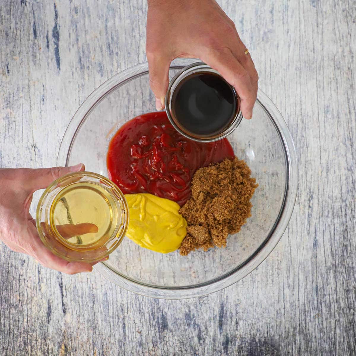 A person holding a small bowl of Worcestershire sauce in one hand and a small bowl of apple cider vinegar in the other hand both over a bowl filled with ketchup, yellow mustard, and brown sugar.
