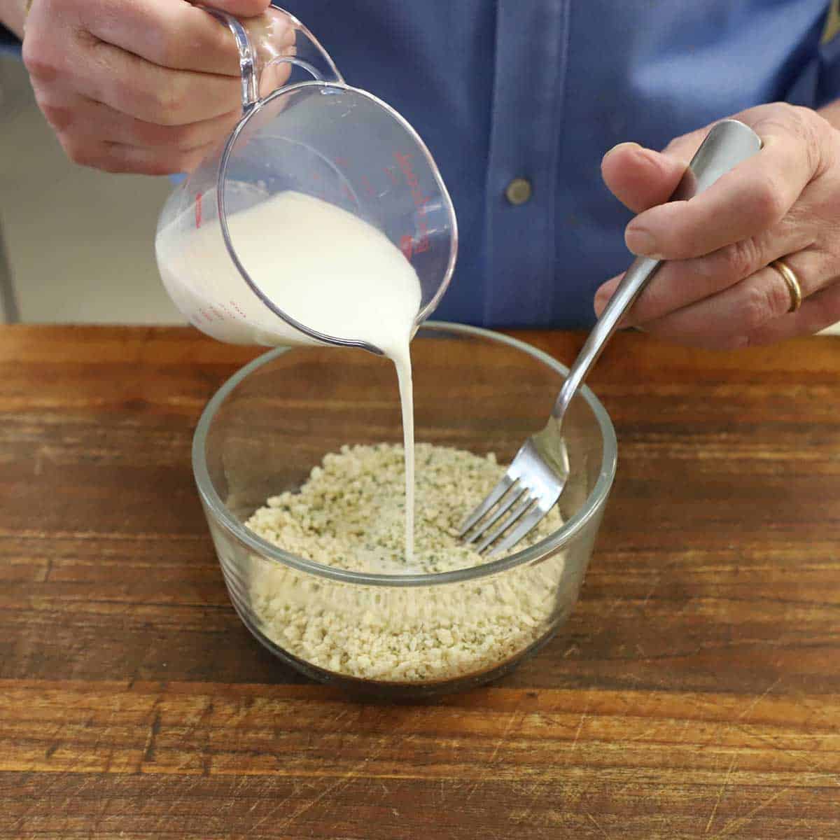 A person pouring whole milk from a small measuring cup into a small glass bowl filled Italian-style Panko breadcrumbs.