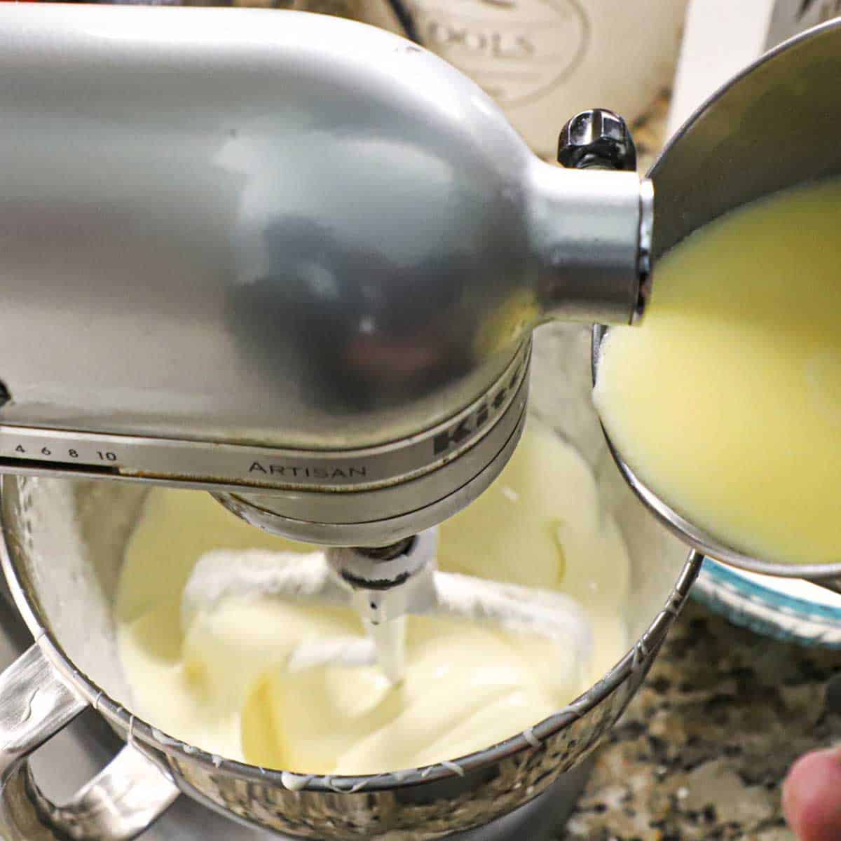 A person pouring warm milk with melted butter from a small saucepan into a the bowl of a stand mixer filled with a batter for custard.