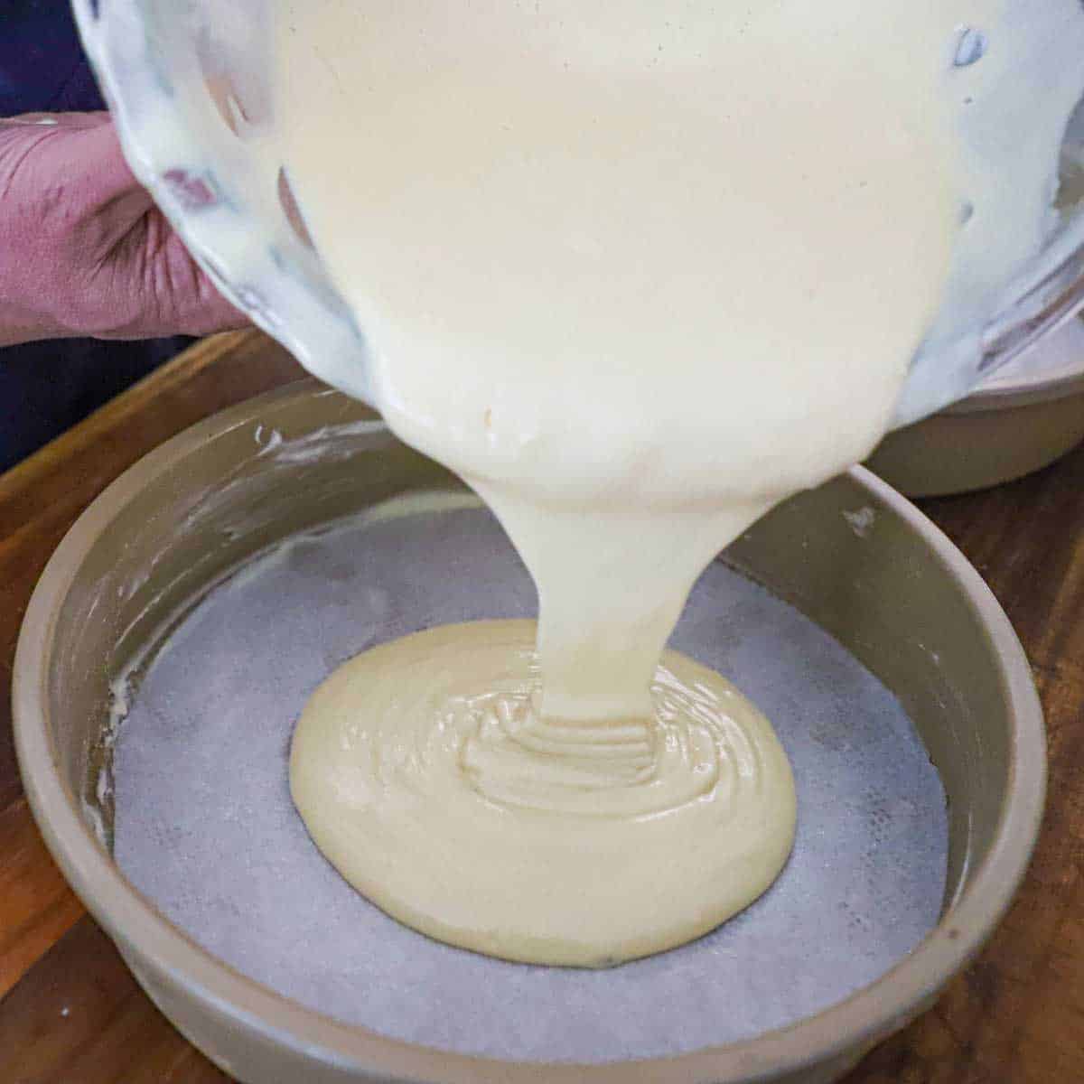 A person pouring a sponge cake batter from a glass bowl into a cake pan that has been greased, floured, and fitted with a circular piece of parchment paper.