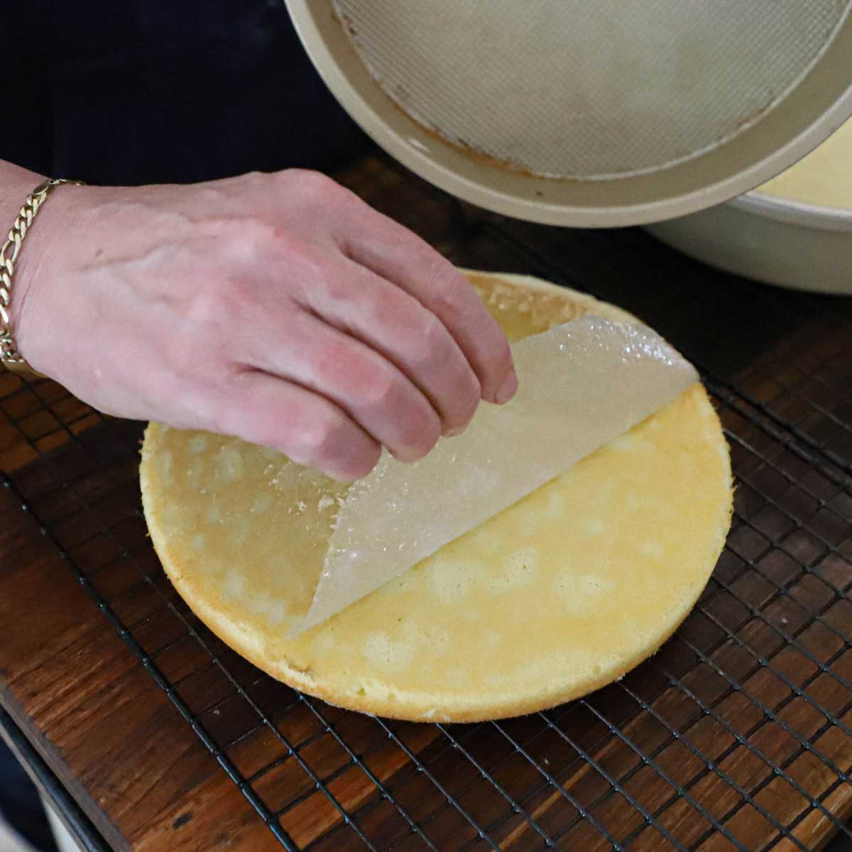 A person pulling a layer of parchment paper from the top of a sponge cake that has been inverted and removed from a cake pan on a baking rack on a wooden cutting board.