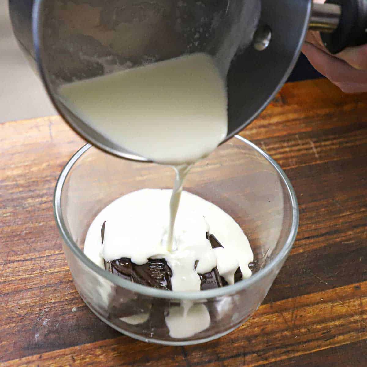 A person pouring hot cream from a saucepan into a small glass bowl filled with chunks of semi-sweet chocolate.
