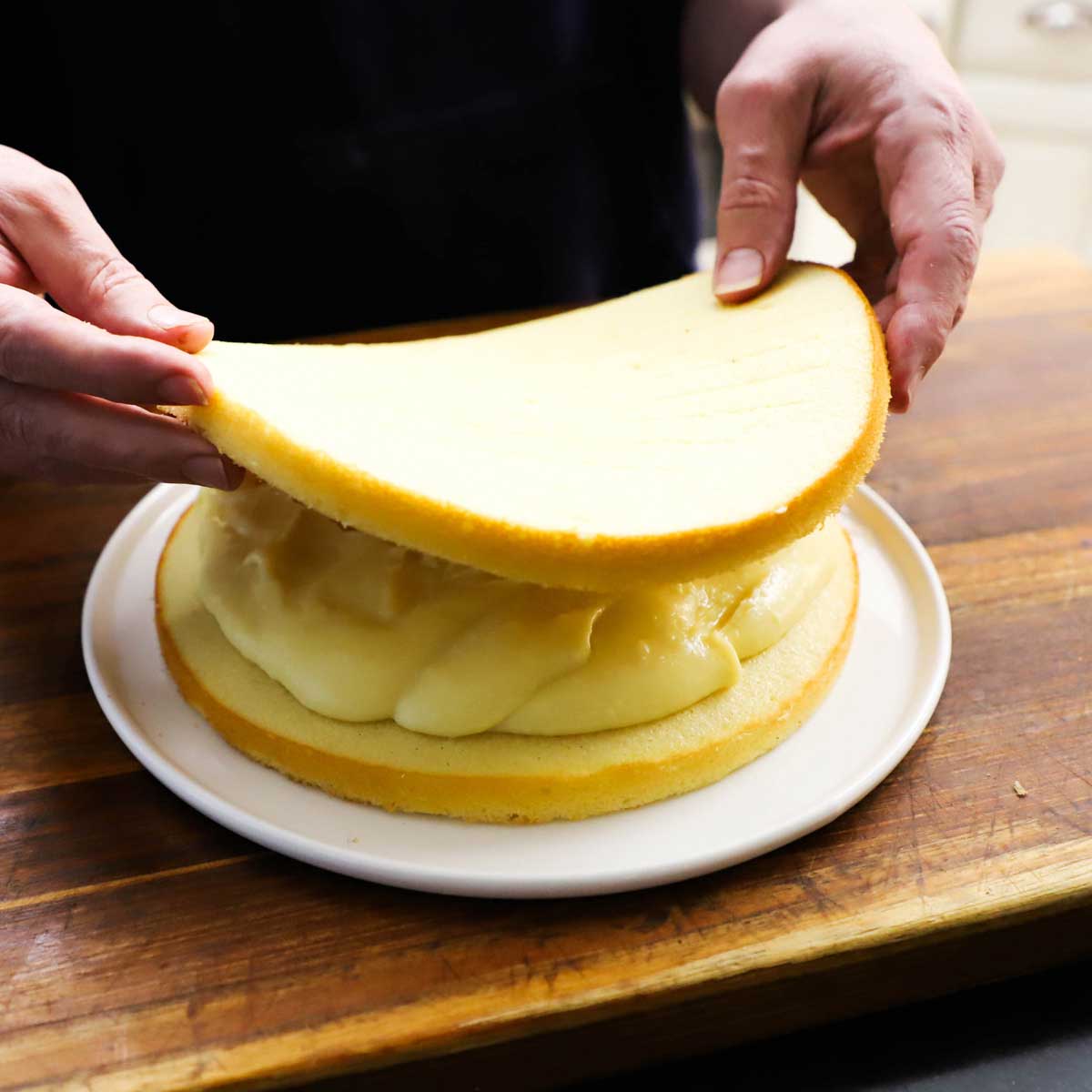 A person placing a thin, circular sponge cake of a mound of vanilla custard that is resting on another circular sponge cake on a white plate on a cutting board.