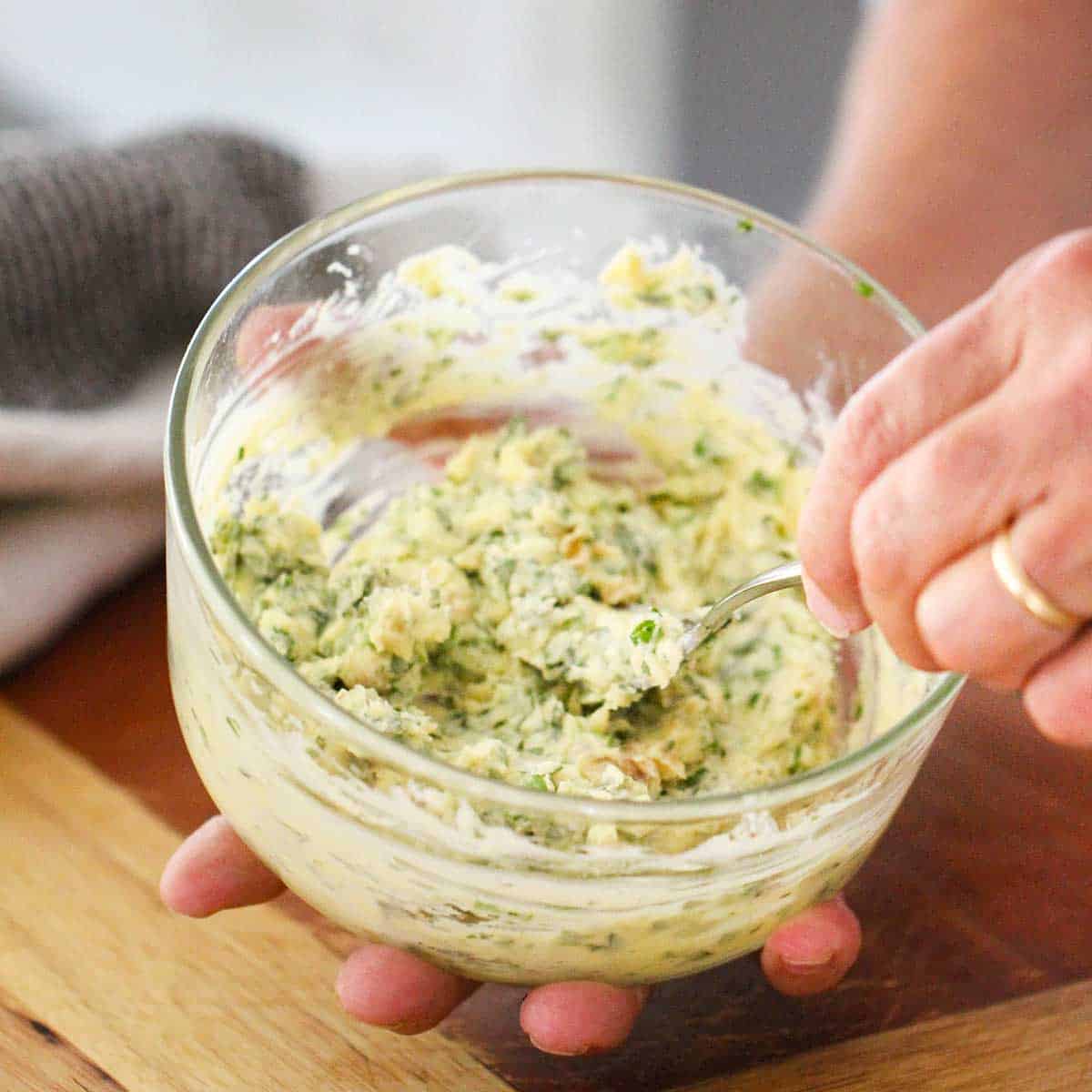 A person using a fork to mash together softened butter, Parmesan cheese, basil, chives, salt, and roasted garlic in a small glass bowl.