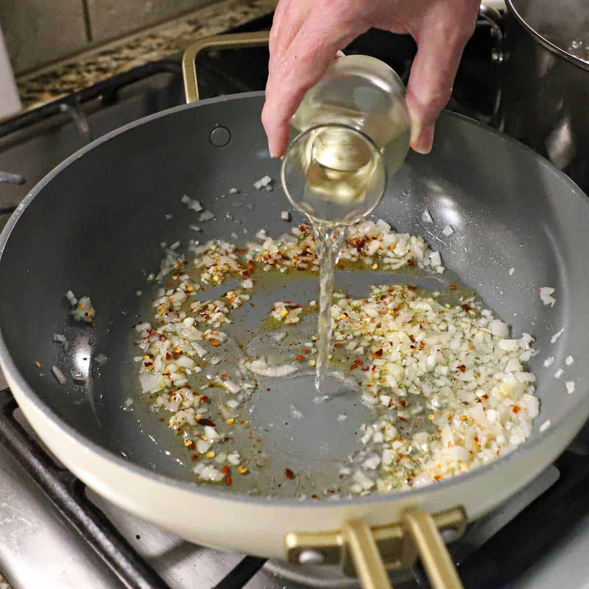 A person pouring white wine from a small glass wine carafe into a skillet filled with sautéed shallots, garlic, and red pepper flakes on a gas stove.