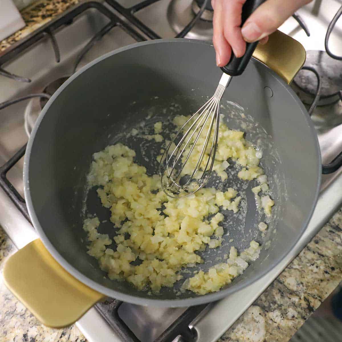 A person using a whisk to stir sautéed onions with flour to create a roux in a large pot on gas stove.