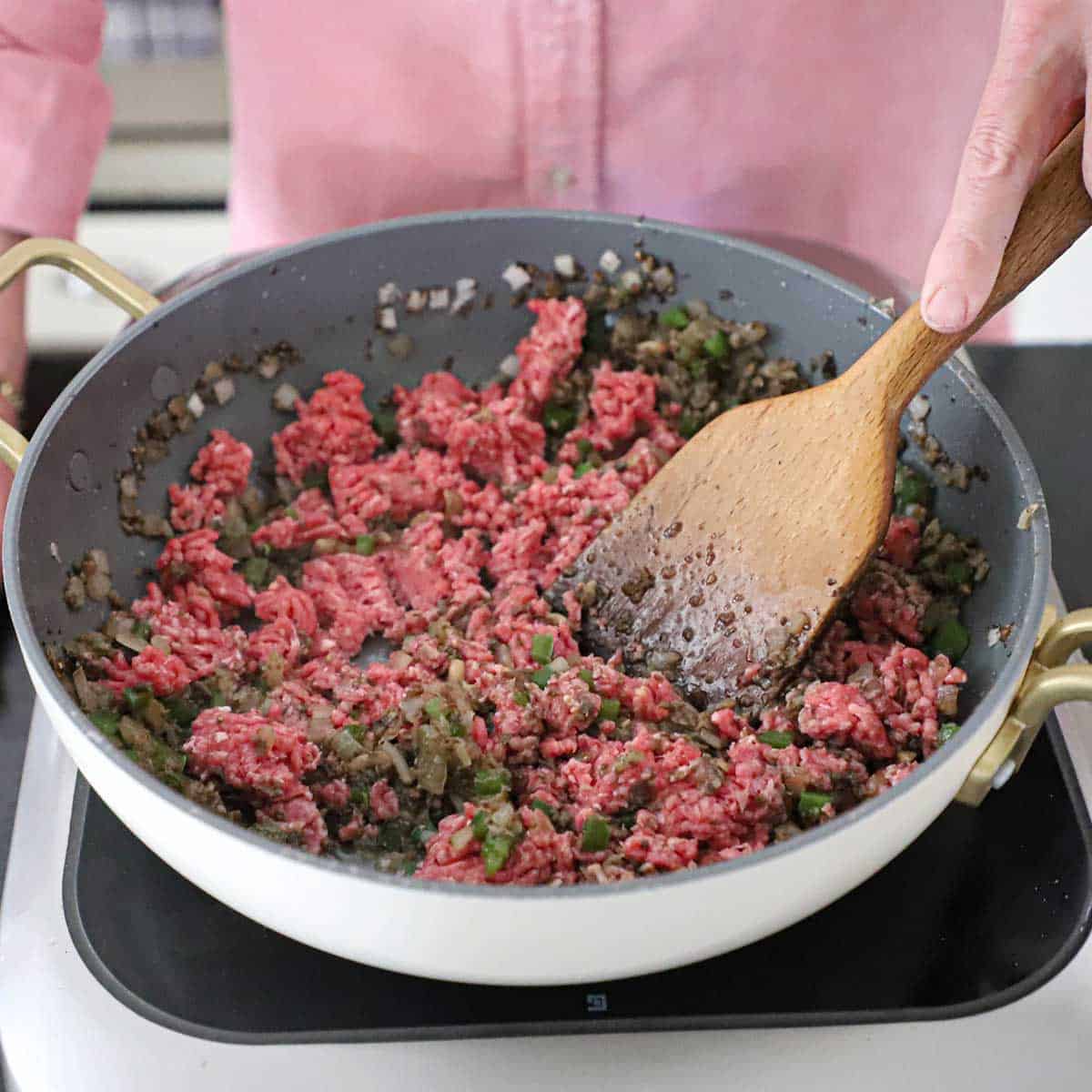 A person using a large wooden spoon to stir ground beef in a hot skillet with sautéed onions, green bell peppers, and chopped mushrooms.