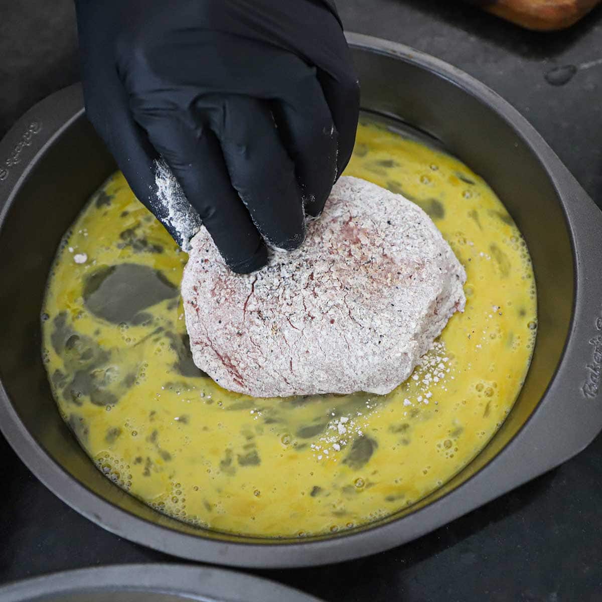 A person dredging a floured boneless pork chop through an egg bath that is in a round baking pan.