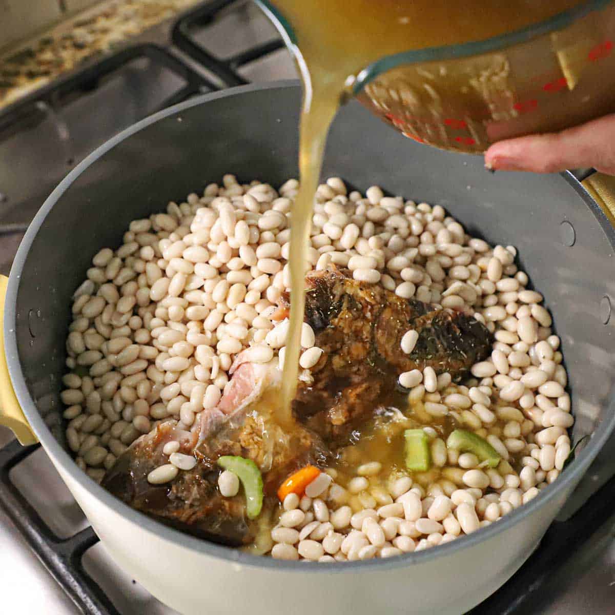 A person pouring homemade chicken stock into a large stock pot that is filled with soaked navy beans, a ham bones, and chopped vegetables.
