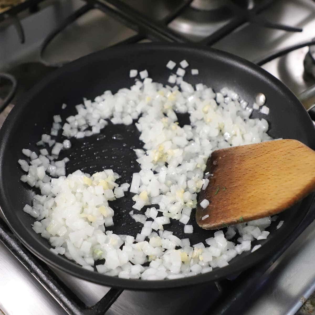 A person using a wooden spatula to stir and sauté chopped onion and minced garlic in a black non-stick skillet over a gas stove.