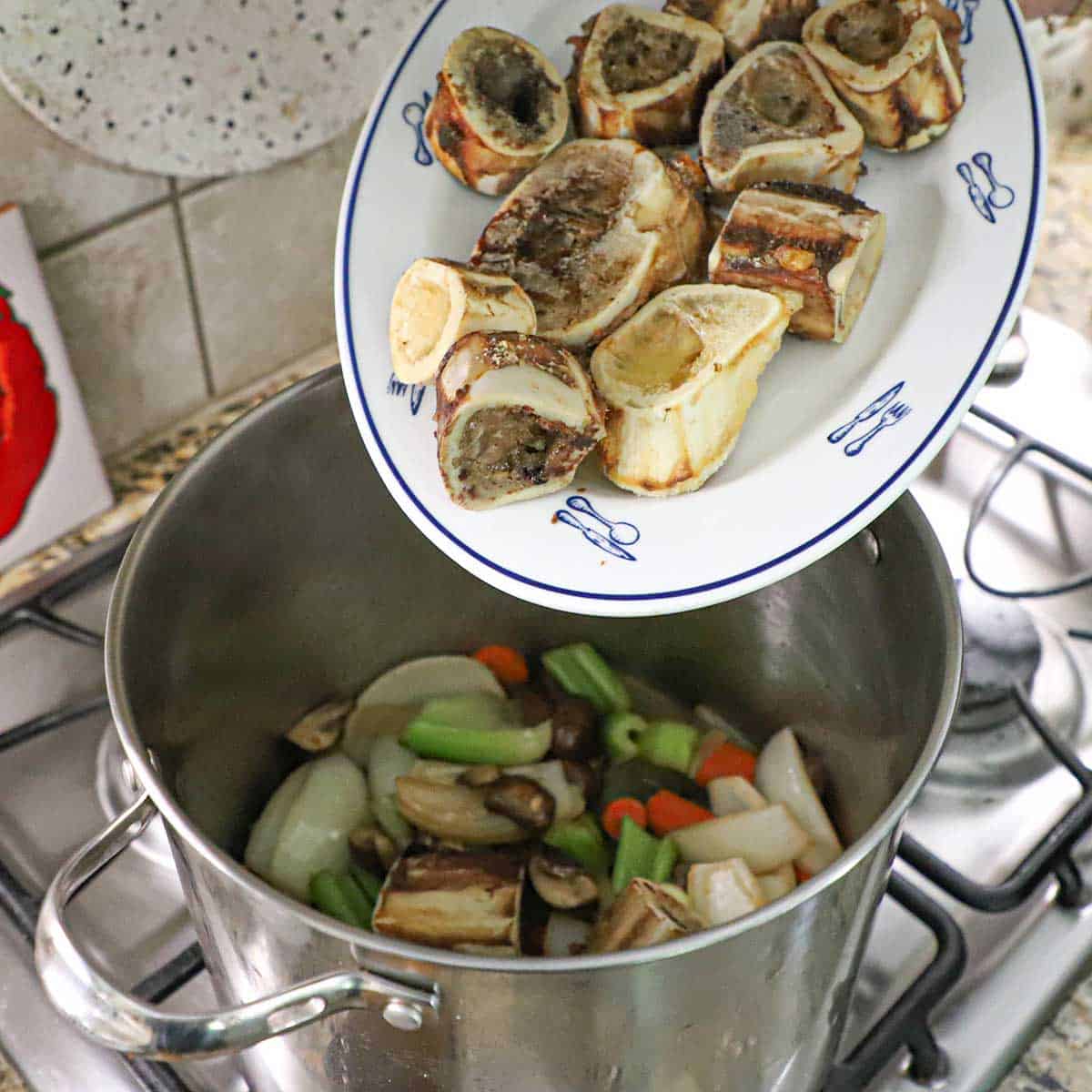 A person transferring roasted beef marrow bones from a large platter into a stock pot filled with sautéed vegetables and mushrooms.