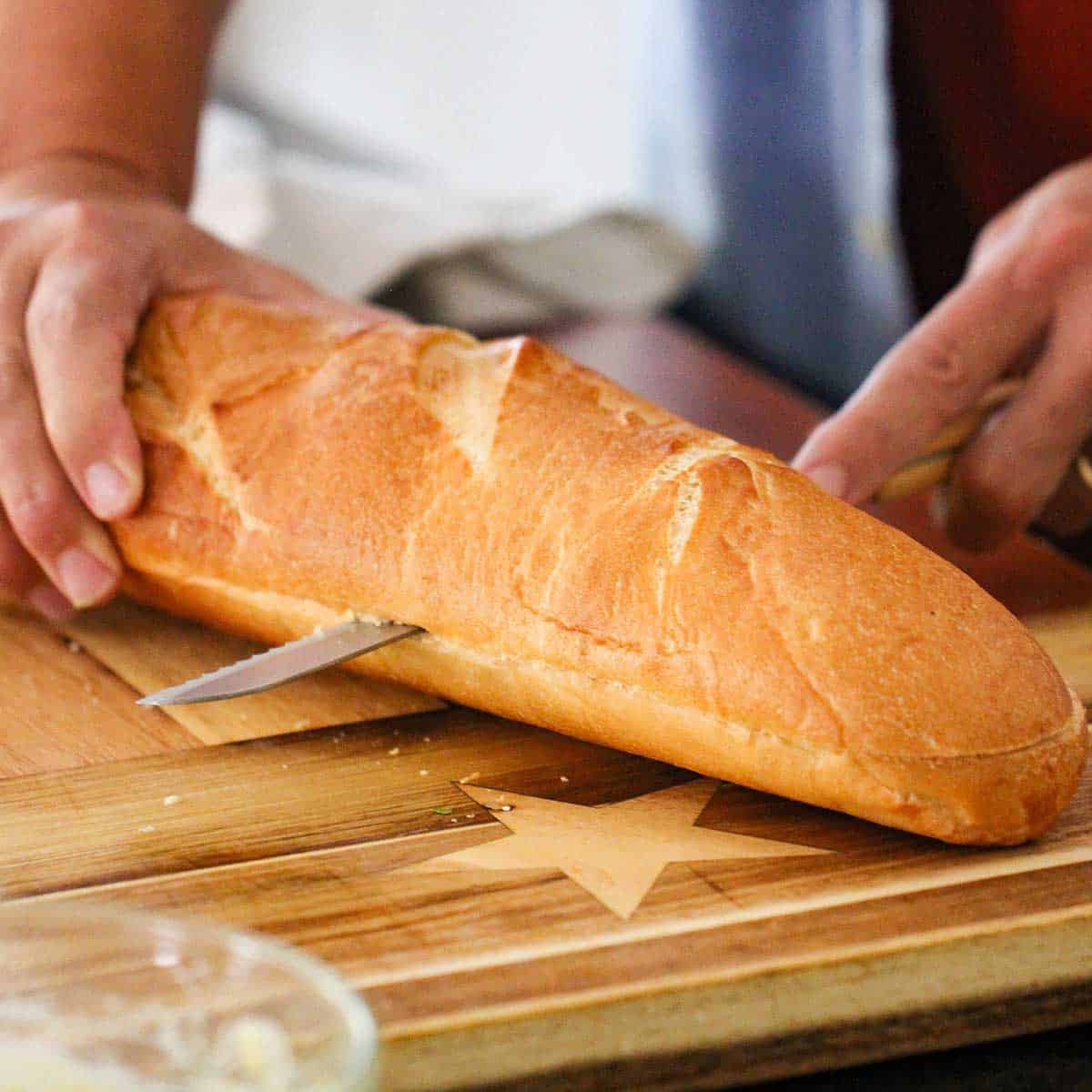 A person using a large serrated knife to cut a large Italian loaf of bread in half, lengthwise on a wooden cutting board.