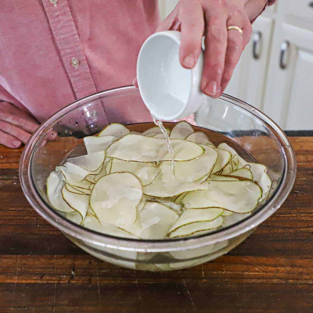 A person pouring distilled white vinegar from a small white bowl into a glass bowl that is filled with thinly sliced potato slices submerged in water.