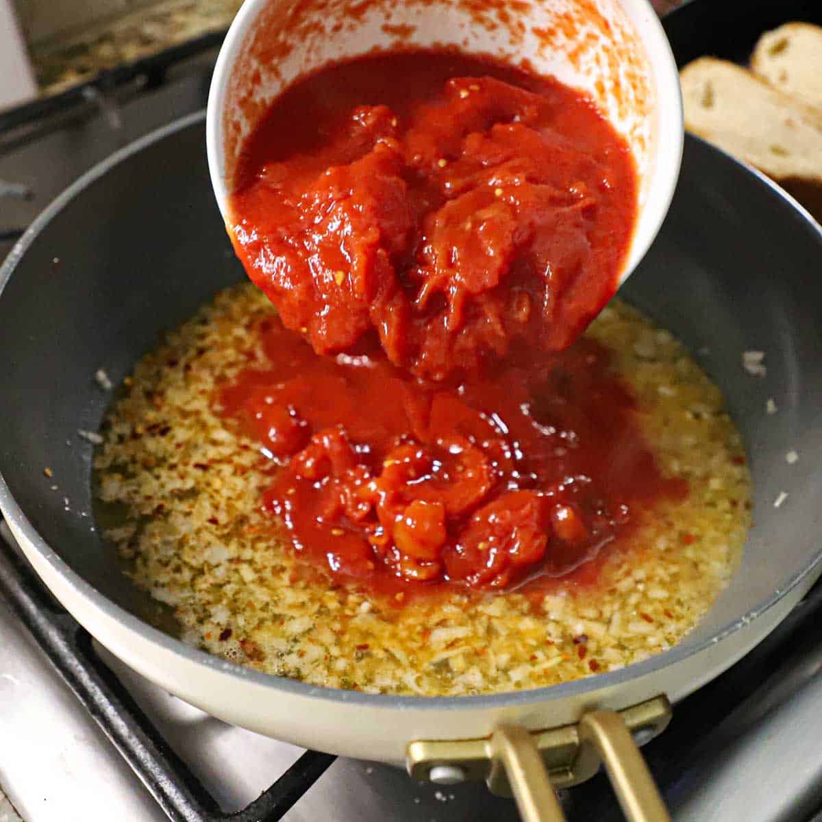 A person transferring hand-crushed whole tomatoes from a white bowl into a skillet filled with a white wine and olive oil sauce with shallots and garlic.