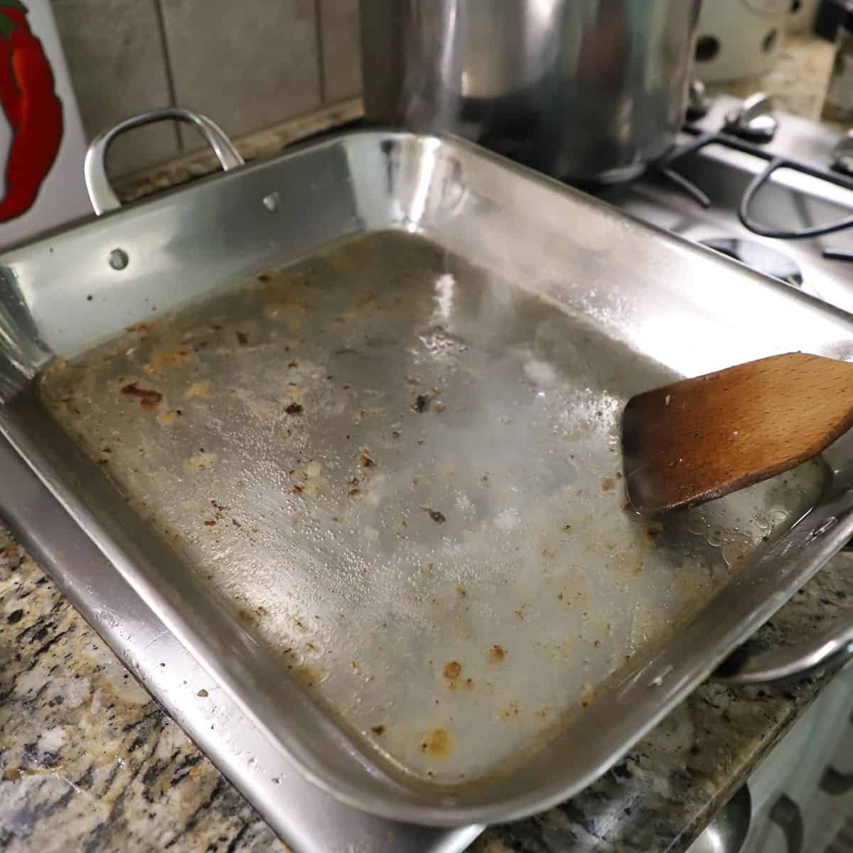 A person using a wooden spatula to deglaze a large steel roasting pan that is being heated on a gas stove across two burners.