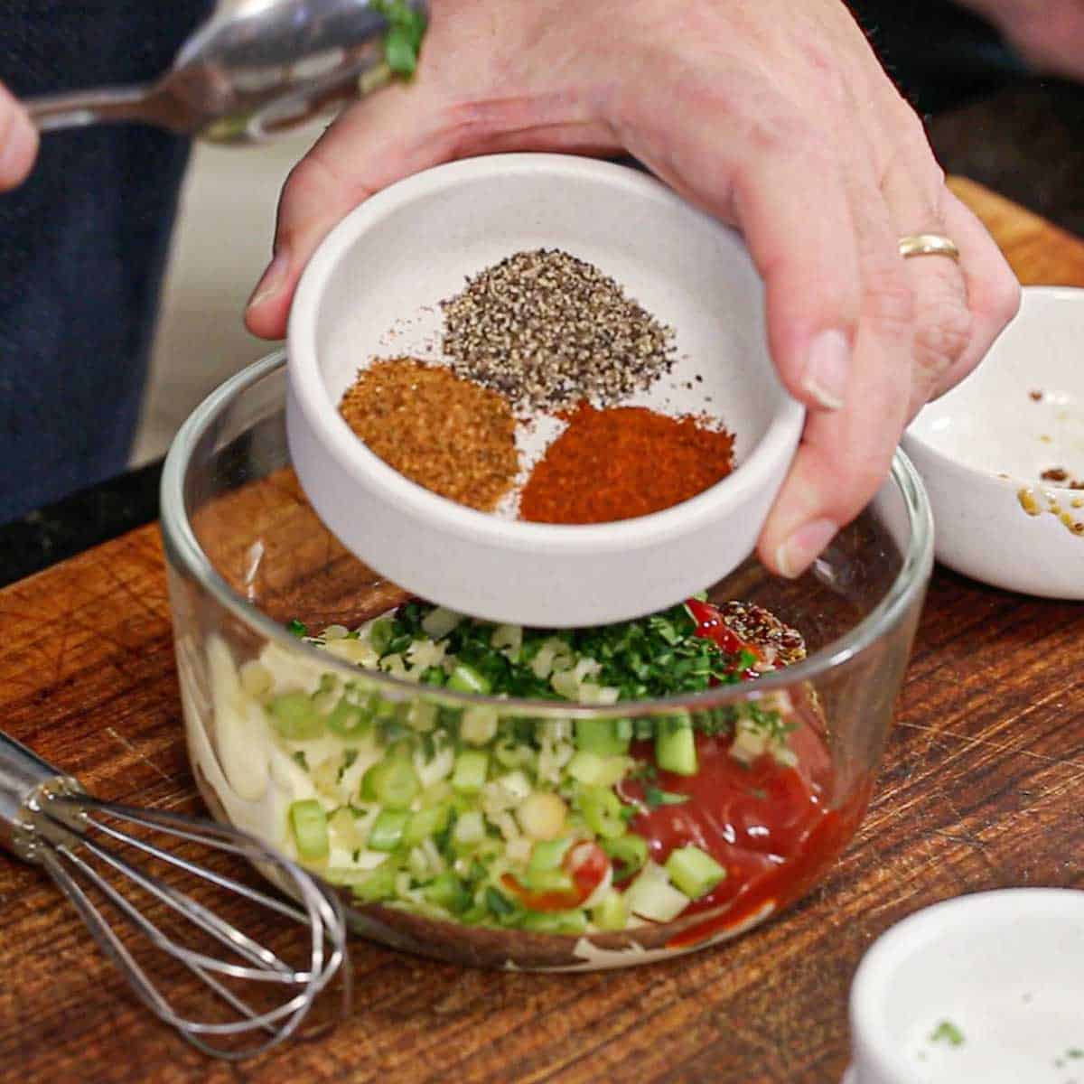 A person holding a small white bowl filled with small piles of black pepper, smoked paprika, and Cajun seasoning over a glass bowl filled Cajun remoulade ingredients.