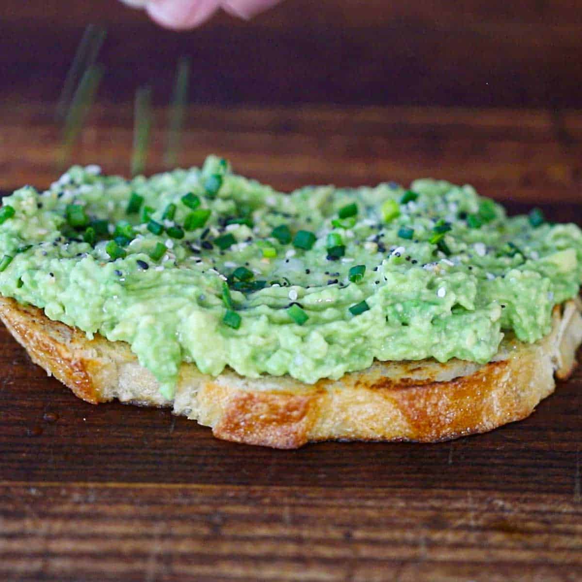 A close-up view of someone sprinkle seasonings over the top of a classic avocado toast on a wooden cutting board.