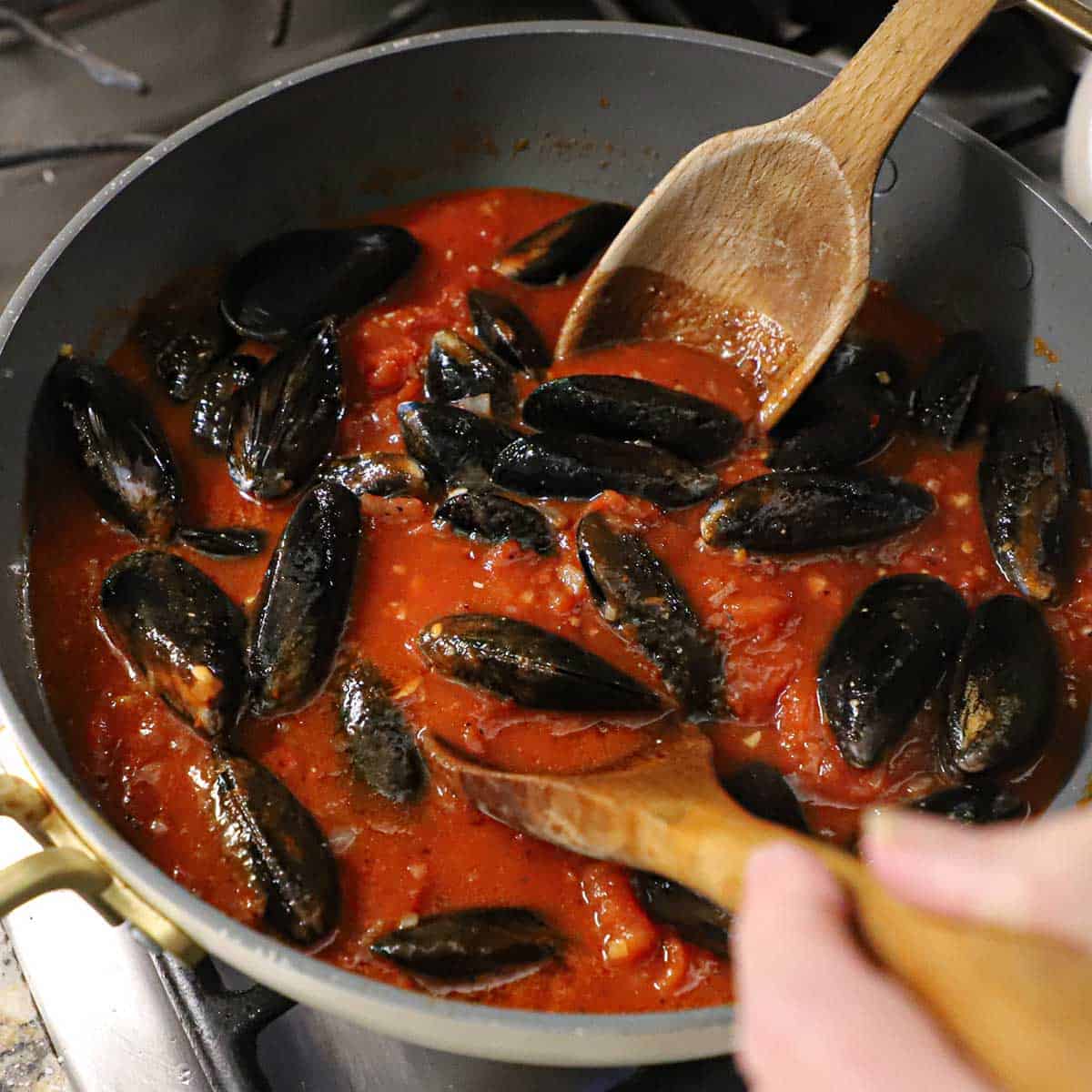 A person using two large wooden spoons to gently toss fresh, unopened mussels in a large skillet filled with a homemade marinara sauce.