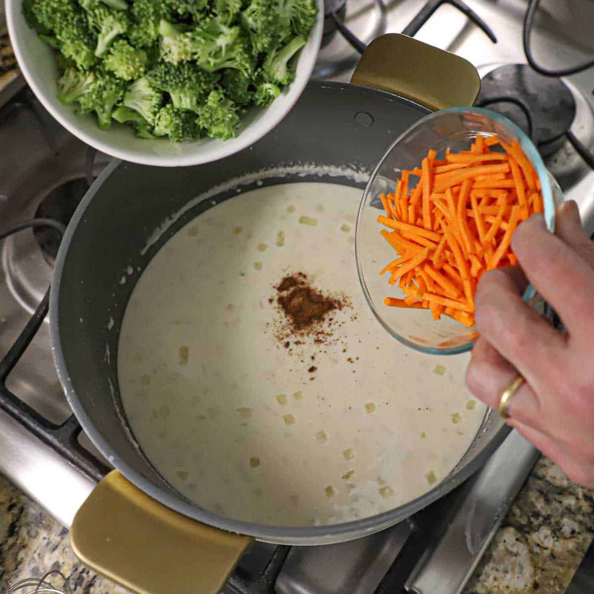 A person holding a small bowl filled with julienned carrots in one hand and another bowl filled with chopped broccoli in the other hand over a bowl filled with a creamy soup in a pot on a gas stove.