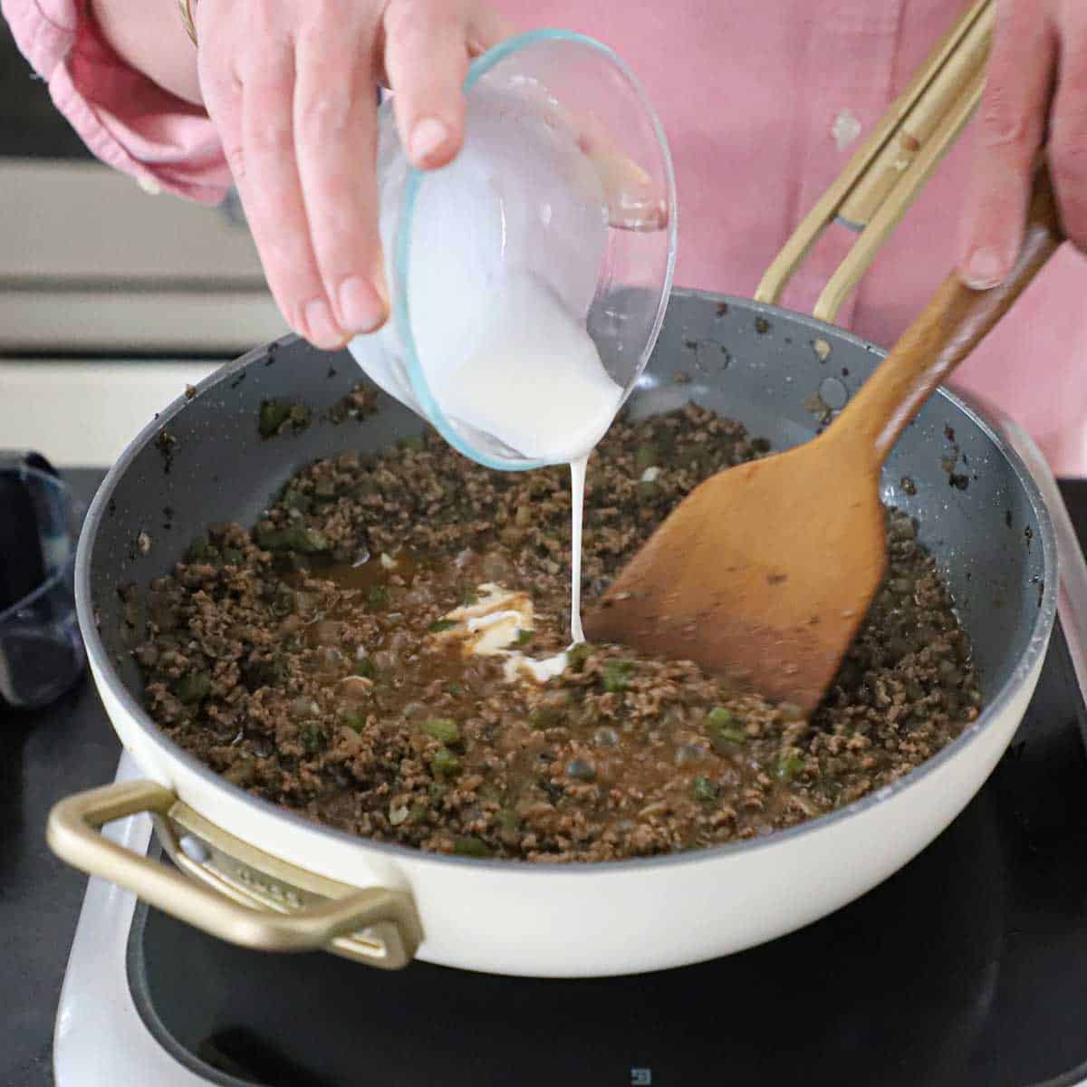 A person pouring a cornstarch slurry into a skillet filled with a simmering Philly cheesesteak sloppy Joes mixture.