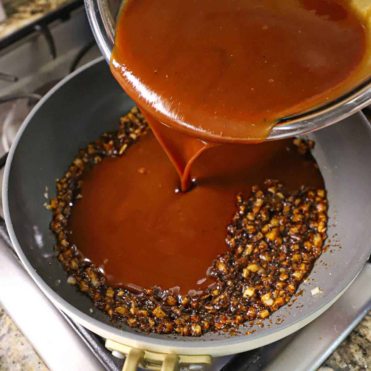 A person pouring barbecue sauce into a large skillet that is filled with sautéed onions and seasonings.